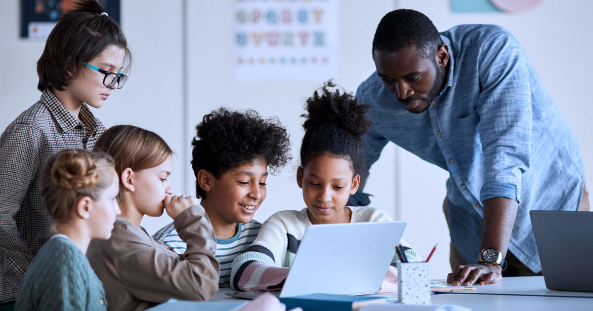A teacher helping a group of students learn from a digital device. 