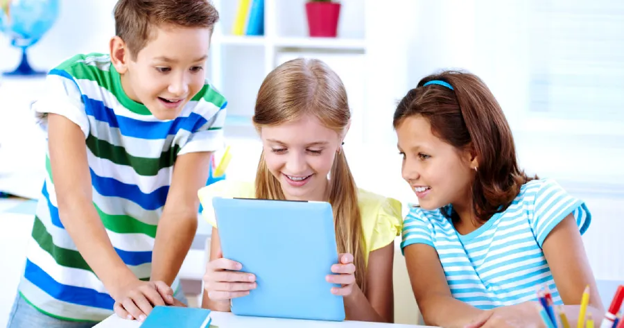 One girl is sitting holding a tablet and one boy and a girl are sitting beside her watching the tablet screen.