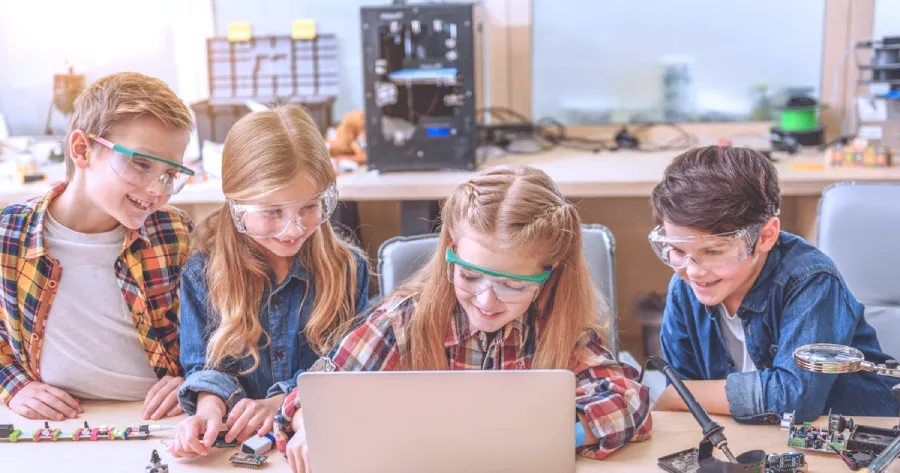 A group of kids wearing safety goggles and sitting in front of a laptop in a classroom. 