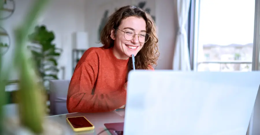 A smiling young woman sitting at her desk utilizes an adaptive learning platform on her laptop. 