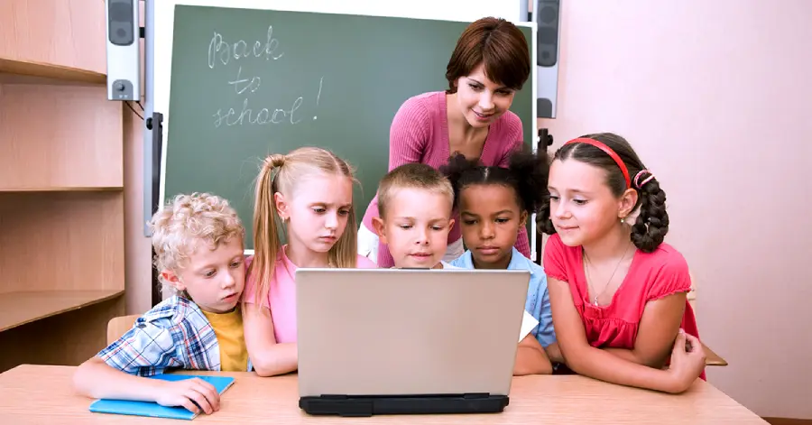 A teacher guides a diverse group of young students as they engage with digital learning material on a laptop, showcasing an early-stage curriculum development in the classroom.