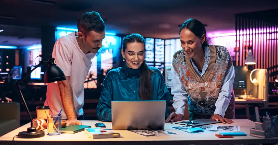 Three people collaborating around a laptop in a modern office environment, reviewing digital content related to curriculum development.
