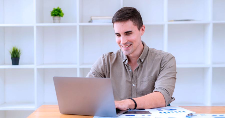 A working professional on a laptop at a desk with printed charts, reviewing the documents online during an ADA website compliance audit in a bright office with white shelving and small potted plants in the background.