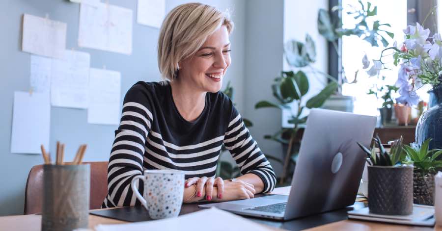A woman working on a laptop in a bright home office with plants and notes on the wall, reflecting the use of AI in education publishing to streamline digital content creation and review.