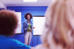 A mentor leads a learning and development session, standing in front of flip charts and explaining a concept to a group of participants in a classroom setting.