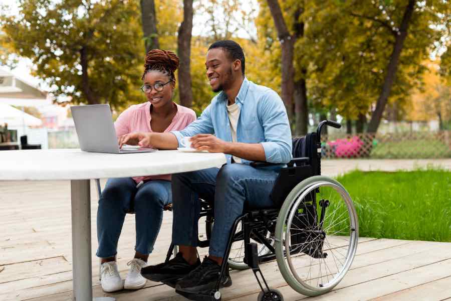An image of two colleagues sitting outdoors at a patio table, reviewing content on a laptop and discussing a microcredential launch plan, with one person using a wheelchair and both smiling in a park setting.