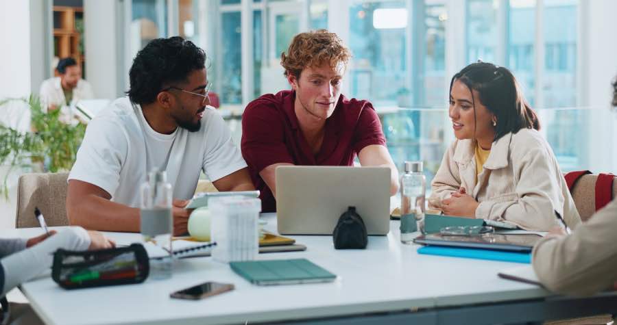 A group of students collaborating around a laptop in a modern classroom, discussing digital content aligned with ADA, WCAG, and VPAT accessibility compliance standards.