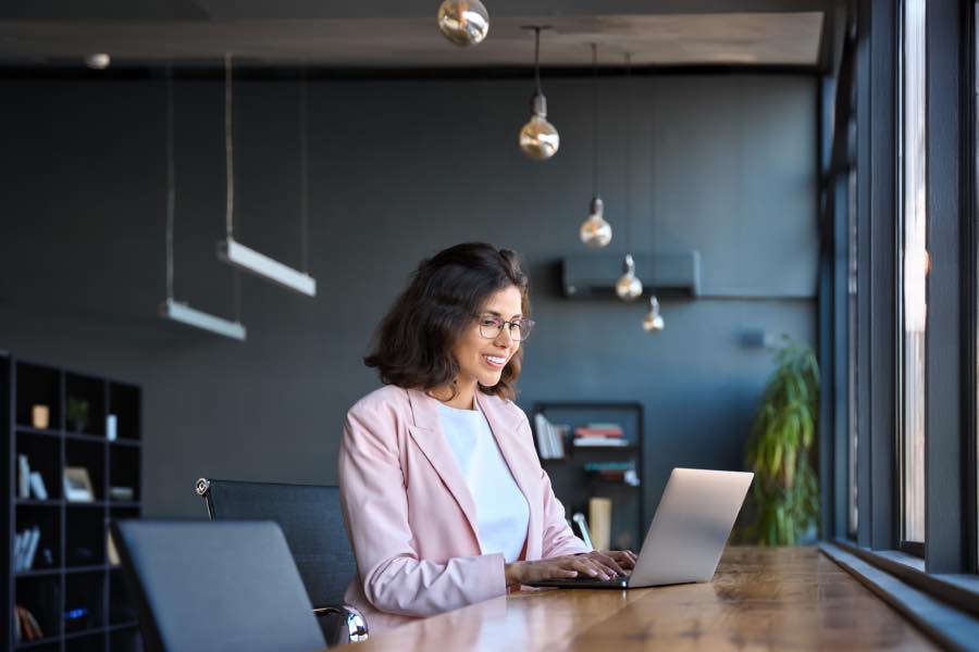 A female working professional on a laptop in a modern office setting, reviewing the digital content for ADA Title II accessibility compliance in an education or publishing workflow.