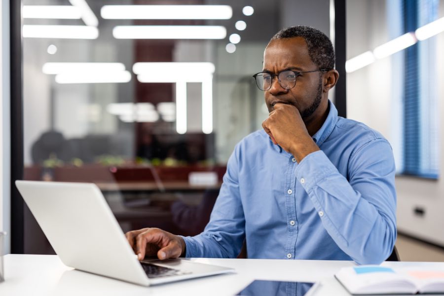 A working professional on a laptop in a modern office, reviewing digital content and identifying issues as part of higher education accessibility remediation at scale, with notes and devices placed on the desk nearby.