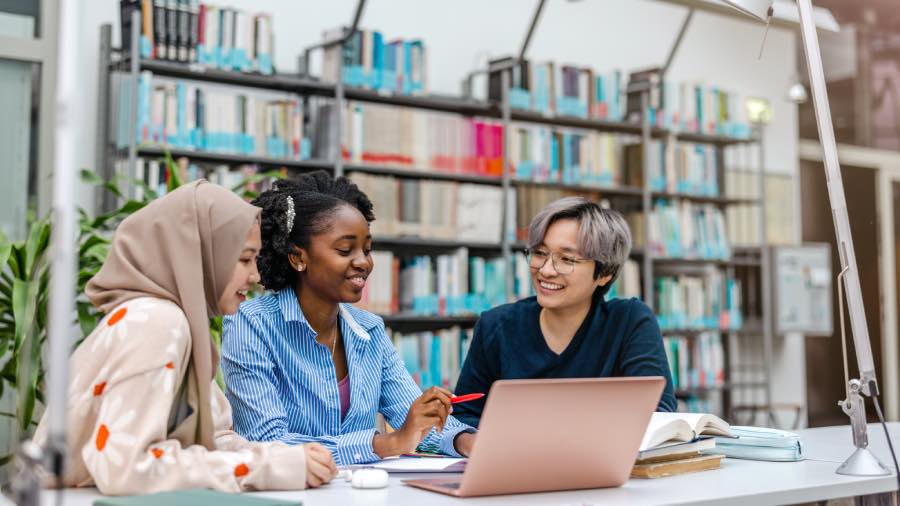 A group of students collaborating around a laptop in a library while reviewing coursework, representing how LMS data analytics for student success can help educators track learning progress and improve outcomes.