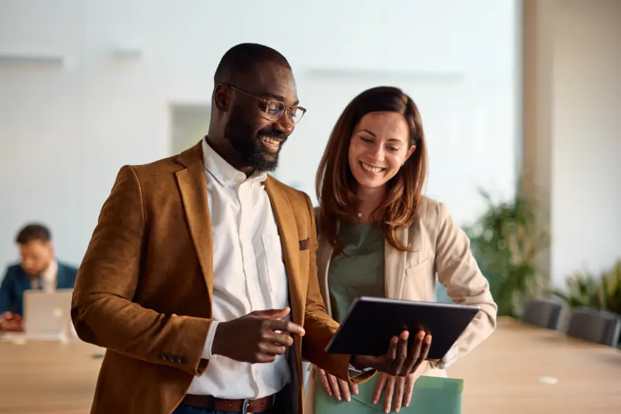 Two coworkers in a contemporary office environment discussing material together using a tablet device, grinning while working on processes for managing learning media, with a blurry background of another coworker in the office.