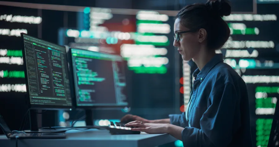 A software developer working across dual monitors displaying code and system data dashboards, highlighting AI-led legacy modernization through the upgrade and maintenance of complex enterprise software systems.