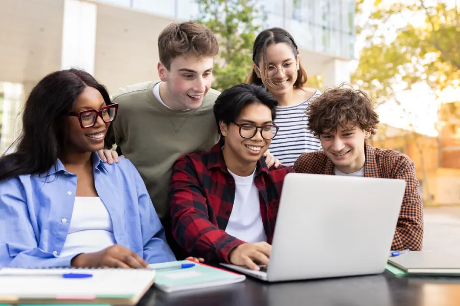 A group of students smiling and learning together on a laptop at a campus table, representing modern digital learning experiences enabled by AI-native content transformation.
