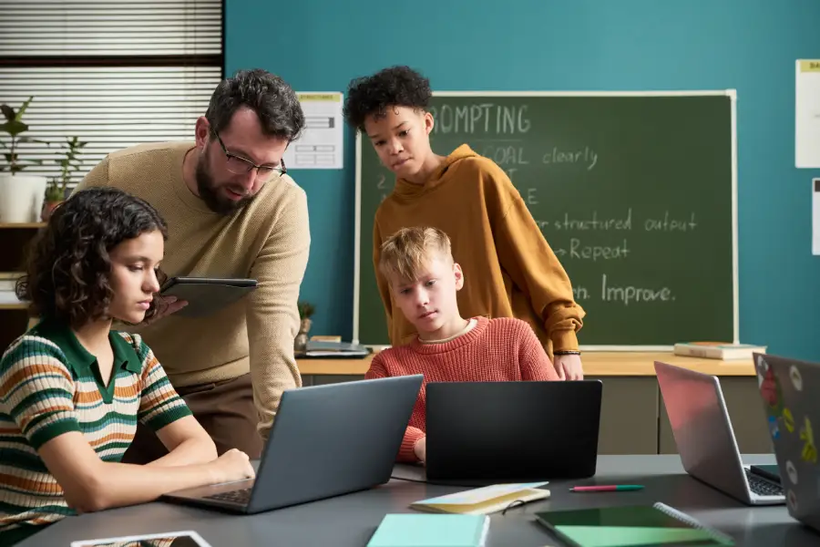 A professor with the students collaborating on laptops in a classroom, reviewing digital learning activities and documentation aligned with the UK edtech procurement requirements, with technology-supported instruction and evaluation in place.