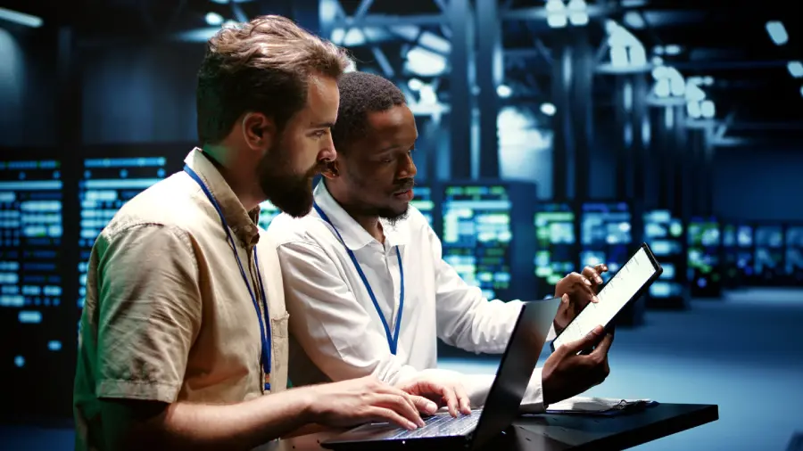 Two working professionals review test data on a laptop and tablet in a server room, demonstrating AI-assisted exploratory testing to identify software issues and validate application performance.