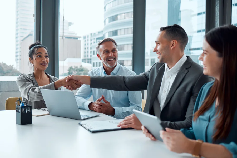 Four professionals in a business meeting, with two professionals shaking hands around a laptop and tablet, showcasing AI in educational publishing partnership.