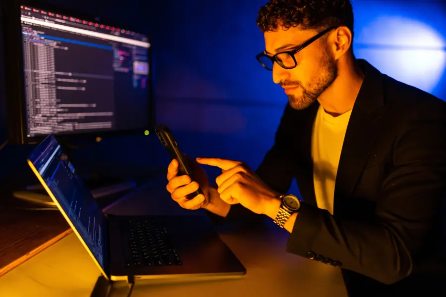 A developer using a smartphone beside a laptop and monitor displaying code as part of an AI-native modernization process.
